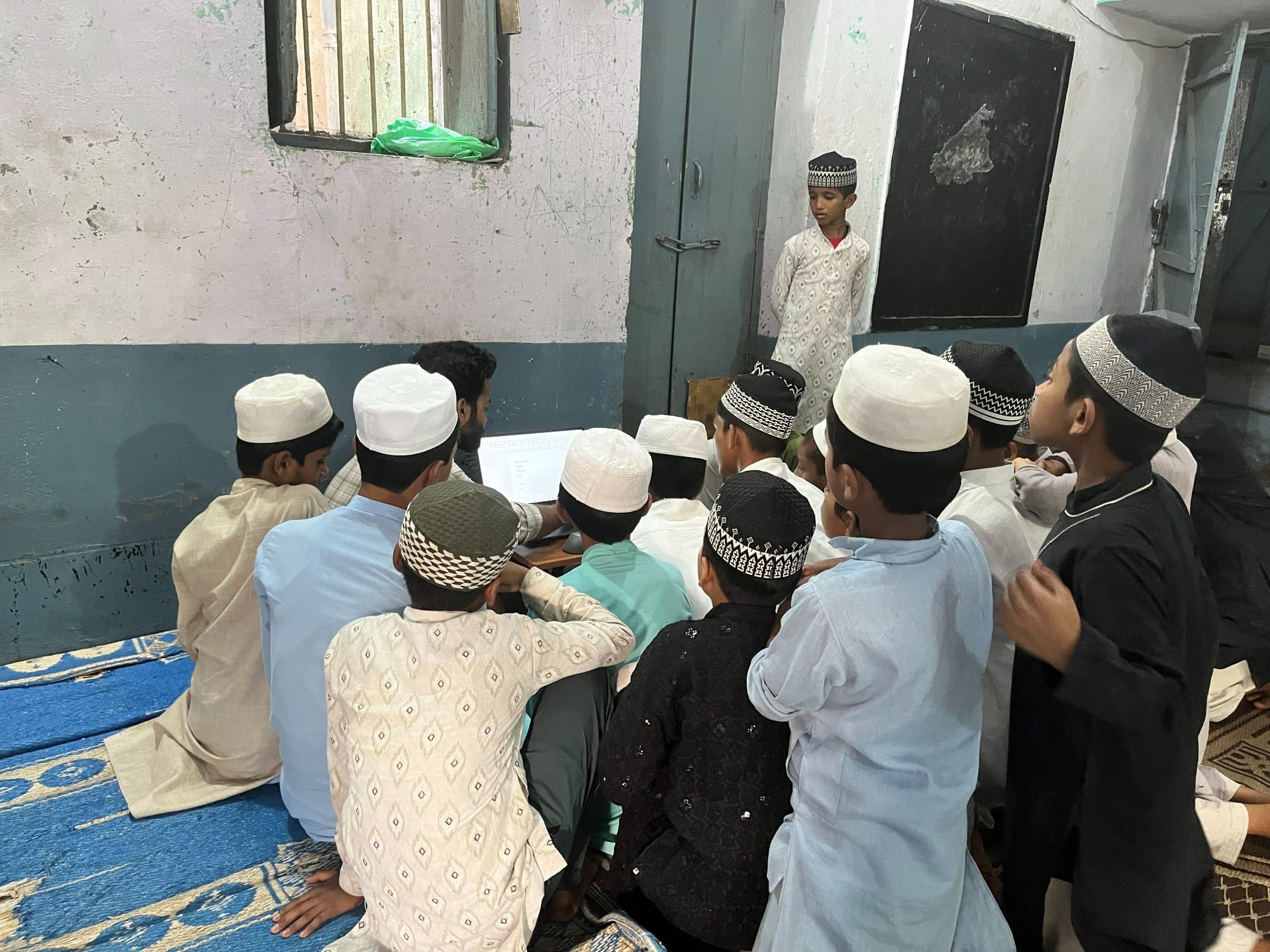 A volunteer-led learning session in a Madarsa classroom, students gathered around a shared laptop.