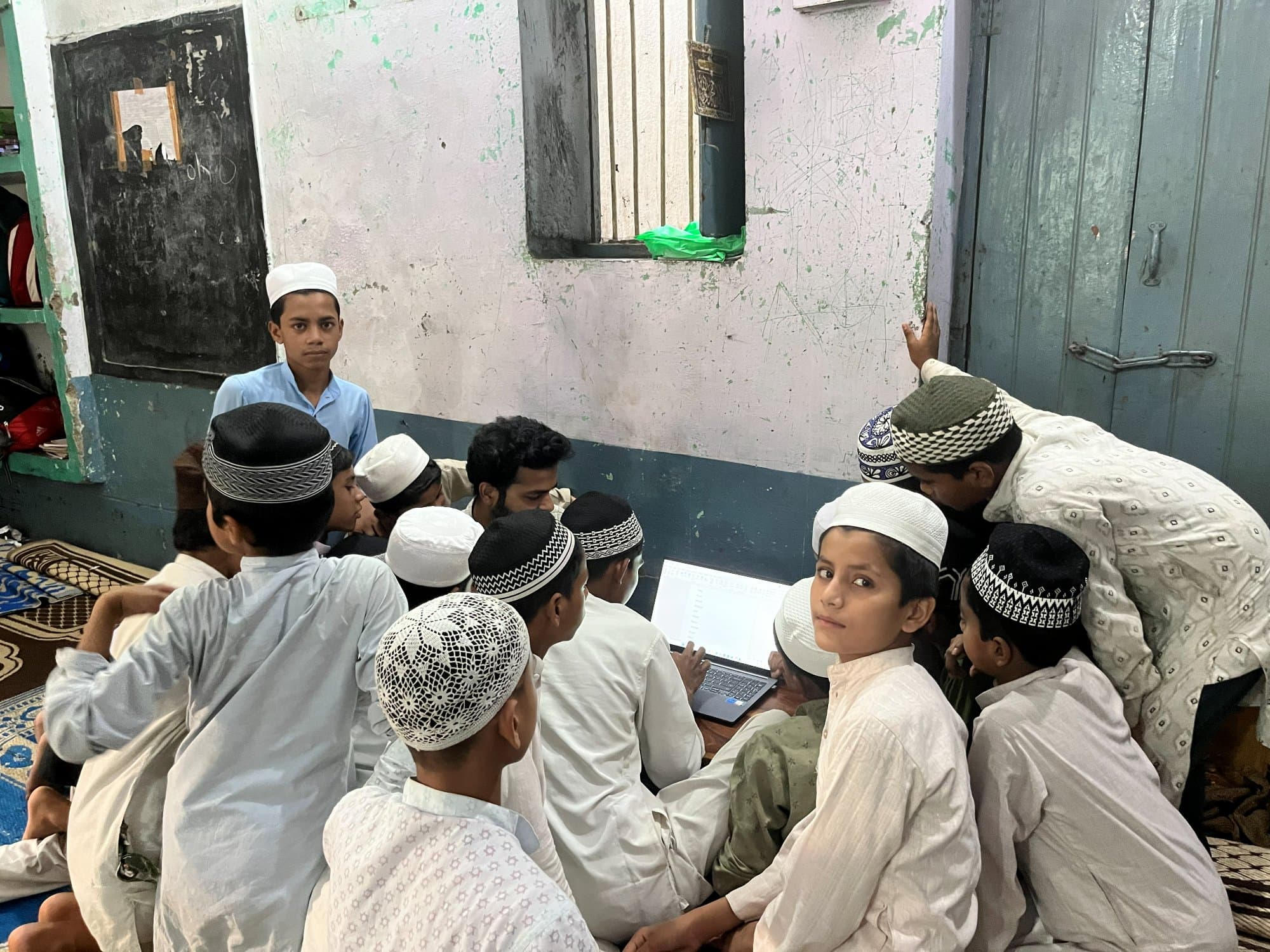 Students crowd around a laptop during an evening digital-literacy session in a Madarsa classroom.